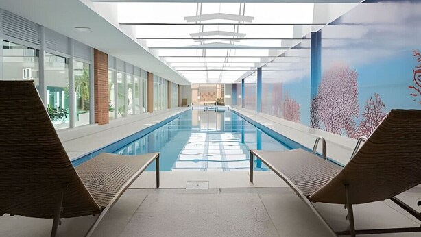 A modern indoor swimming pool with clear glass walls, lounge chairs, and a mural of coral reefs. - Apartamento com Vista do 22º Andar, Piscina Aquecida e Garagem | Batel, Curitiba (Curitiba)
