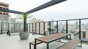 A rooftop terrace with a wooden table and benches, a large planter with a palm tree, and a cityscape view through large windows.