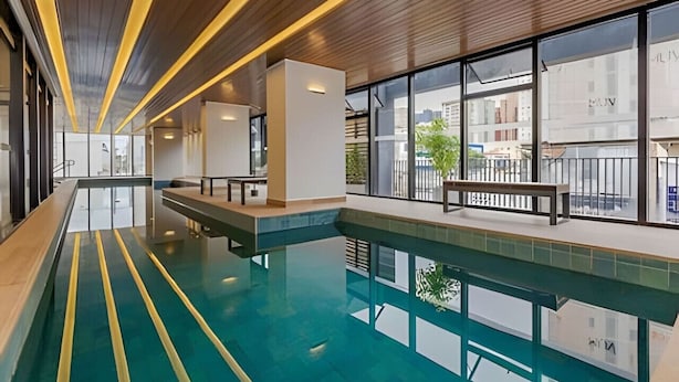 A modern indoor swimming pool with clear glass walls, wooden ceiling, and a bench along the edge.