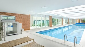 A modern indoor swimming pool with clear glass walls, a brick wall, and a poolside seating area.