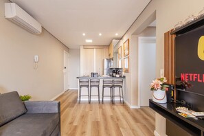 Una sala de estar moderna con un sofá gris, piso de madera, una cocina con barra y un televisor montado en la pared mostrando el logotipo de Netflix.