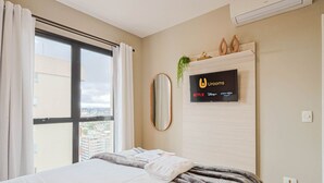 Ein Hotelzimmer mit einem Bett, einem großen Fenster mit Vorhängen, einem angebrachten Fernseher, der Streaming-Dienste anzeigt, und einer kleinen Pflanze an der Wand.