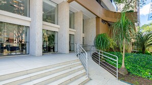 Ein moderner Gebäudeeingang mit einer Treppe, die zu einer Glastür führt, umgeben von Pflanzen und Metallgeländern.