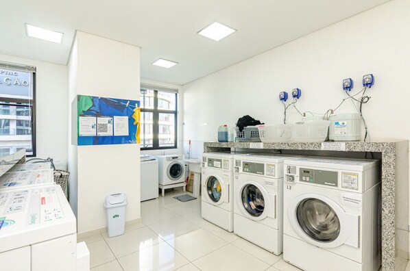 Un cuarto de lavandería con varias lavadoras, un área de secado y un tablón de anuncios con carteles informativos.