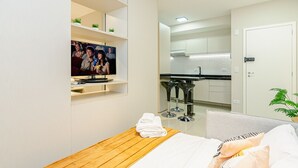 A compact hotel room with a bed, a wooden table with towels, a TV mounted on the wall, a kitchenette, and a door with a handle. - Apto Completo com 1 quarto em frente shop estação. (Curitiba)