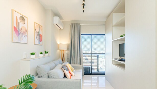 A modern living room with a grey sofa, a small wooden table, and a TV on a white shelf. There are two wall-mounted air conditioners and a window with a view of buildings. - Apto Completo com 1 quarto em frente shop estação. (Curitiba)