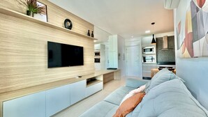 A modern living room with a flat-screen TV mounted on a wooden wall, a grey sofa with a throw pillow, and a kitchen area in the background.