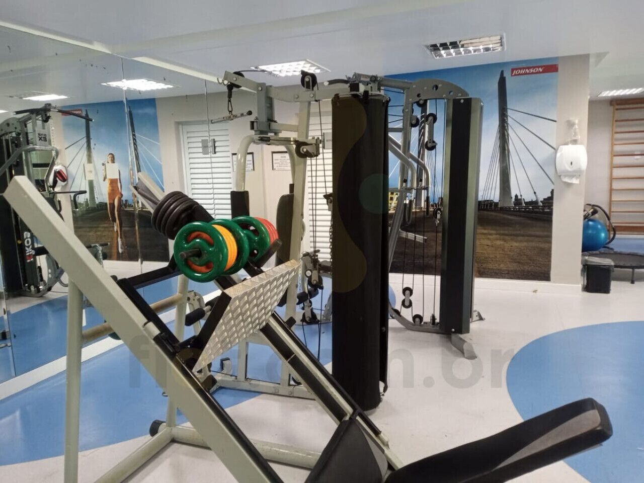 A gym with weightlifting equipment, a blue floor, and a wall with a bridge picture.