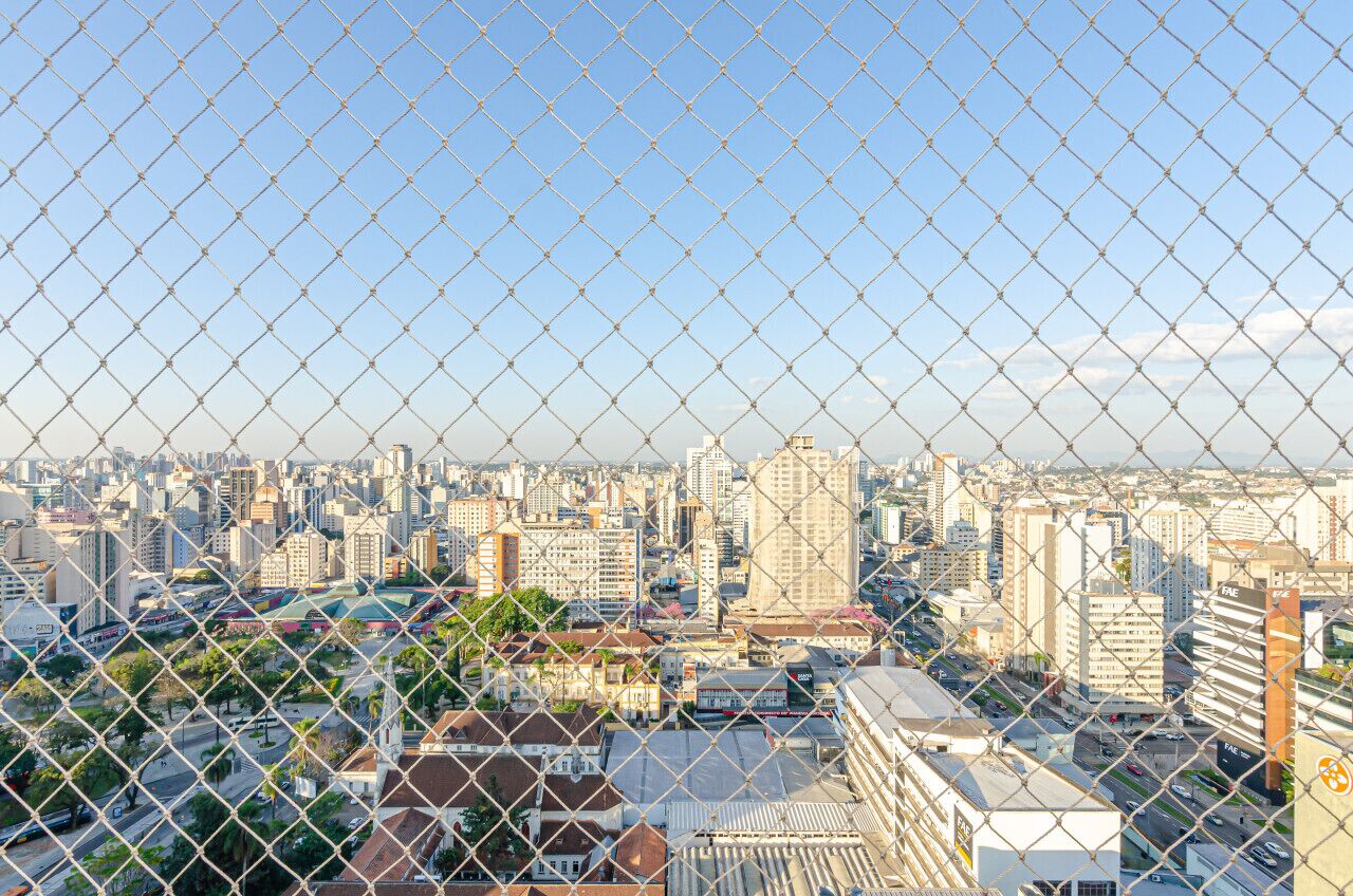 Vista de un paisaje urbano a través de una cerca de alambre, con una mezcla de edificios altos y estructuras más pequeñas.