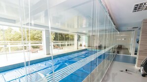 A glass-enclosed indoor swimming pool with clear glass walls, a blue pool floor, and a view of the outdoors. - Studio 25º Andar Sacada e Linda Vista no Hydepark (Curitiba)