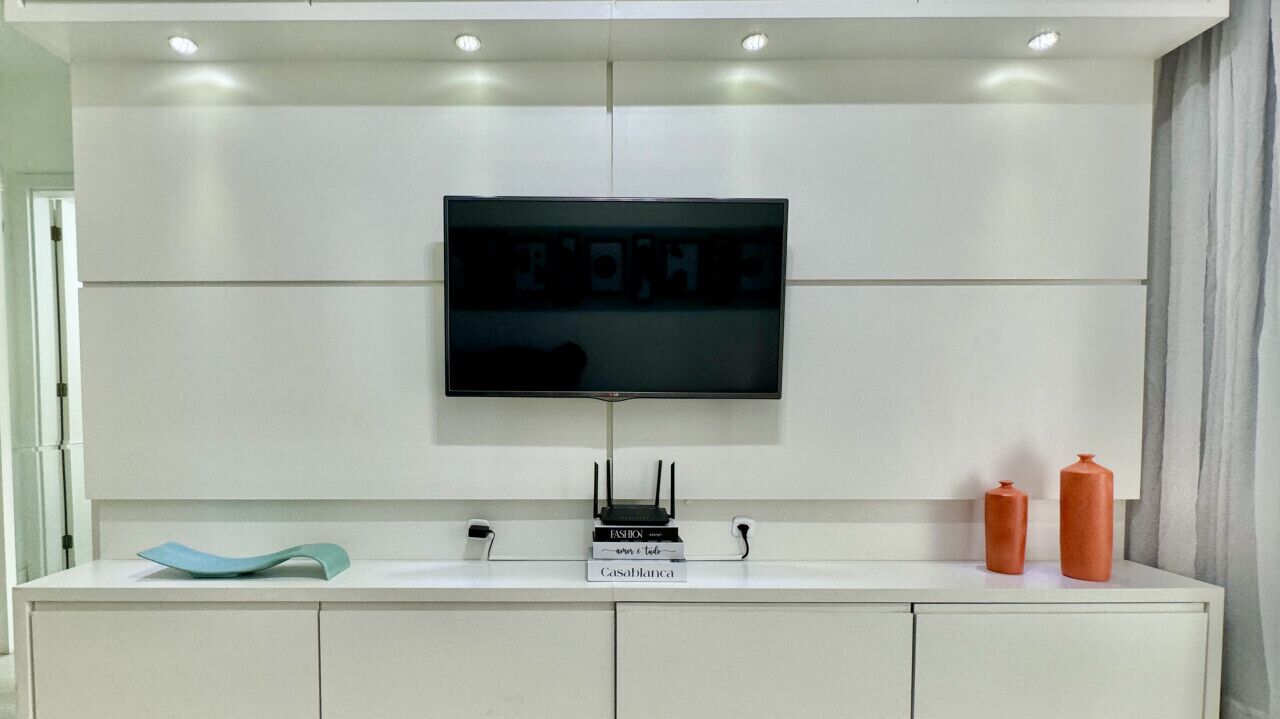 A modern bathroom with a large mirror, a flat-screen TV mounted above the mirror, and a white vanity with a sink and two orange bottles.