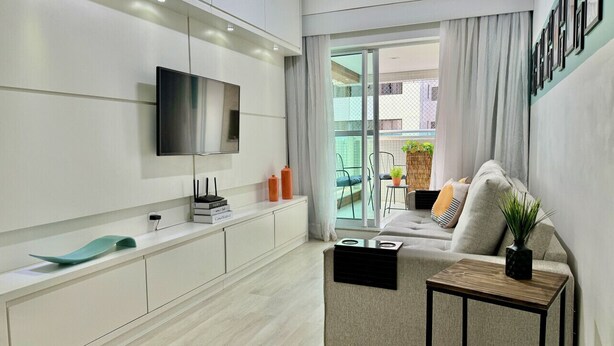 A modern living room with a flat-screen TV mounted on the wall, a sofa with cushions, a small wooden side table, and a sliding glass door leading to an outdoor area.
