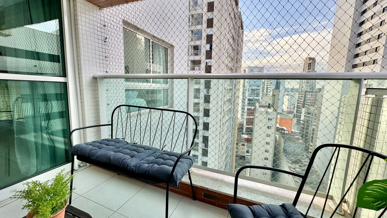 A balcony with a cushioned bench and chairs, a potted plant, and a view of city buildings.