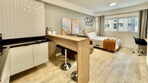 A modern hotel room with a kitchenette, a bed with a white comforter, a large window with curtains, and a view of the city. - Studio Lindo, Condomínio Clube Lifespace Curitiba (Curitiba)