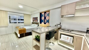 A compact hotel room with a bed, a small kitchenette, a dining area, and a large window.