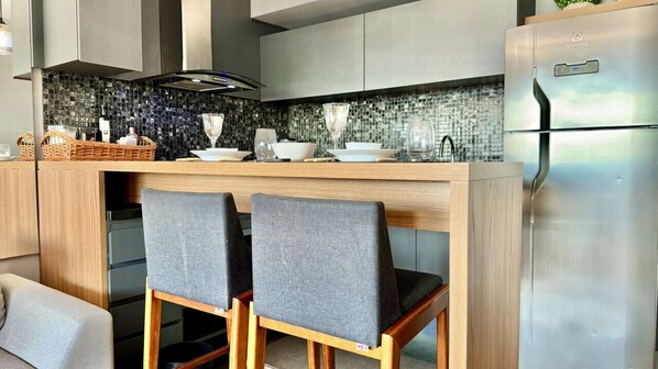 A modern kitchen with a central island, two grey upholstered chairs, a stainless steel refrigerator, and a mosaic backsplash.