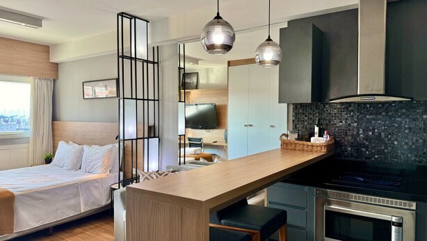 A modern hotel room with a bed, a wooden desk, a kitchenette, and a TV.