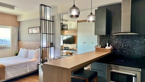 A modern hotel room with a bed, a wooden desk, a kitchenette, and a TV.