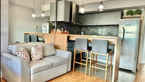 A modern kitchen with a central island, a dining area with bar stools, and a living room with a sofa and decorative pillows.