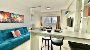 A modern living room with a teal sofa, a small kitchen area with a white countertop, and a large window offering a city view.