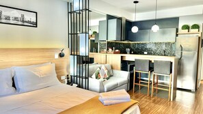 A modern hotel room with a bed, a sofa, a small kitchen area, and a dining table.