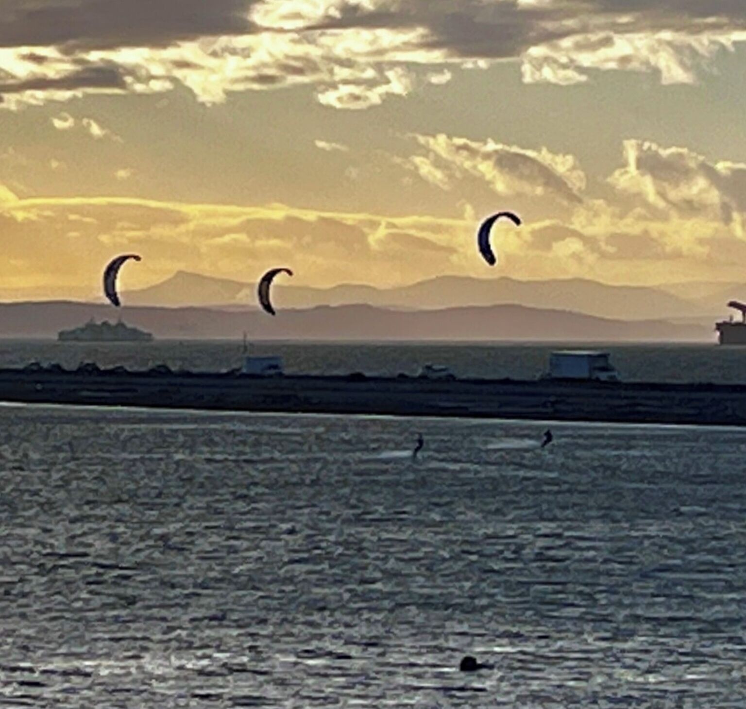 Cena costeira com kitesurf, um navio e montanhas ao longe.