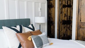 A neatly made bed with white linens and multiple pillows, a bedside lamp, and a wooden door in the background.