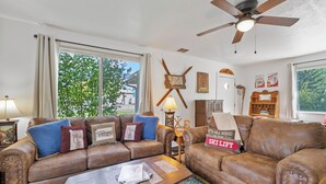 A living room with two brown leather sofas, a wooden coffee table, and a ceiling fan. - 30-Day Min, Walking Distance to Downtown & Lake + Pet Friendly! (Whitefish)