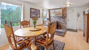 A dining area with a round wooden table, wooden chairs with cushioned seats, a vase with flowers, and a stone wall with a painting. - 30-Day Min, Walking Distance to Downtown & Lake + Pet Friendly! (Whitefish)