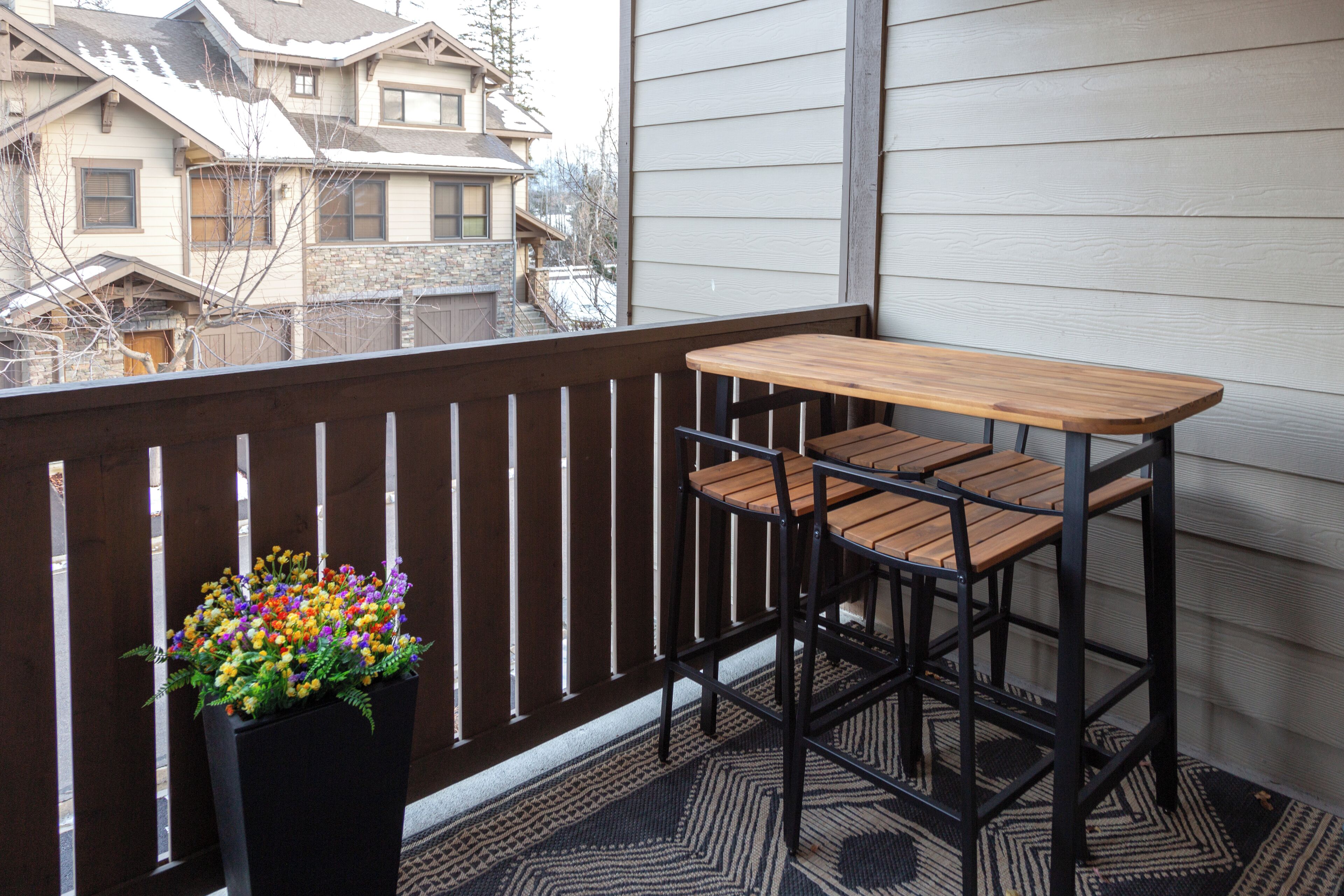 Een balkon met een houten tafel en twee stoelen, een plant in pot met kleurrijke bloemen, en een uitzicht op huizen op de achtergrond.