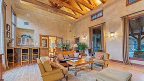 A spacious living room with wooden floors, a large sofa, a coffee table, and a fireplace. There are wooden chairs and a small table with a lamp. The room has a high ceiling with exposed beams and large windows.
