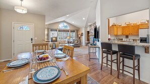 A spacious dining area with a wooden table set for a meal, a kitchen with wooden cabinets, and a large window providing natural light. - 30-Day Min, Close to Downtown, Walk to Golf & Lake! (Whitefish)