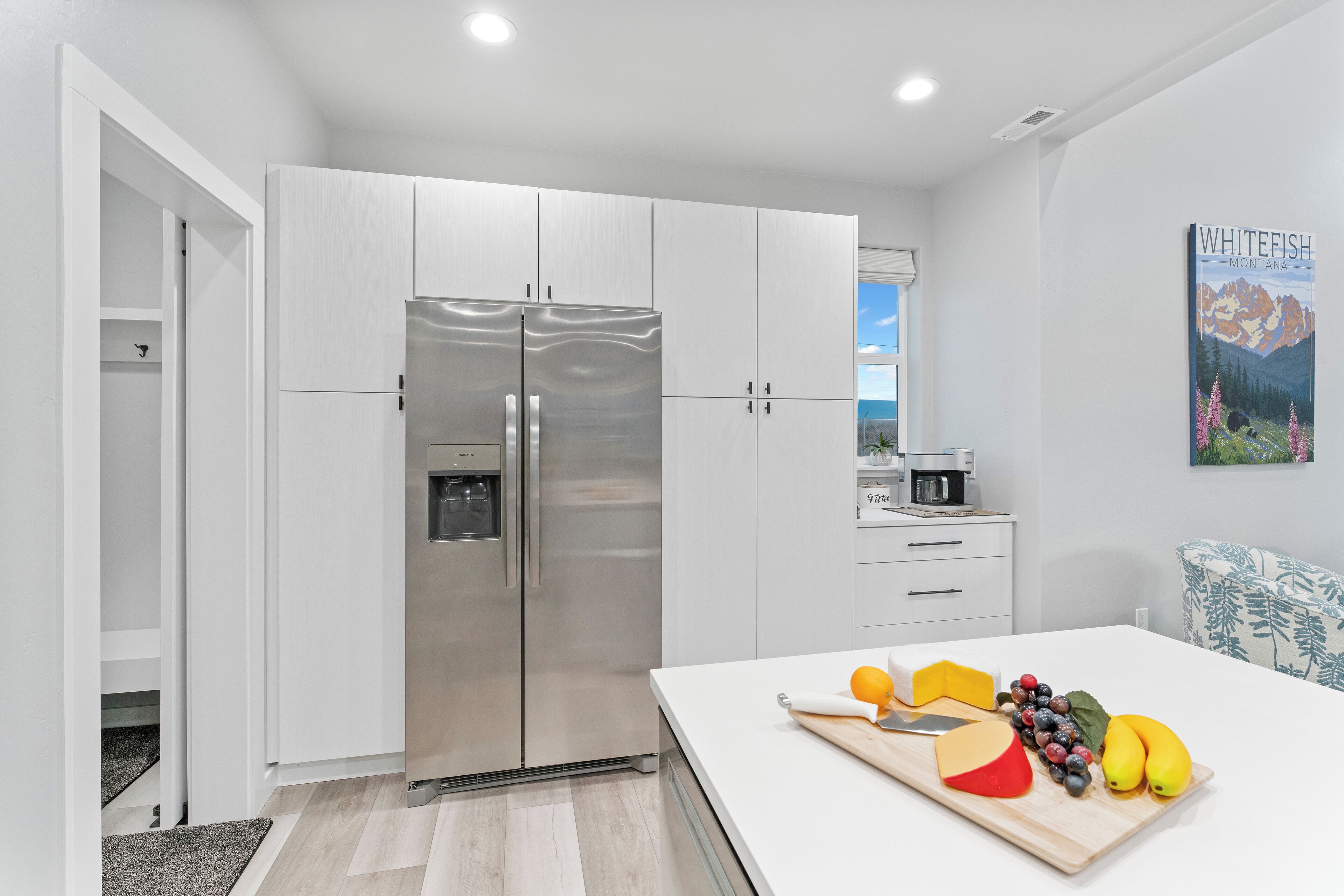 A modern kitchen with white cabinetry, a stainless steel refrigerator, and a dining area with a table set with fruit and cheese.