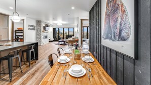 A modern dining area with a long wooden table set for a meal, a stone fireplace, and a large bear painting on the wall. - Hot Tub + Close to Downtown, Smith Fields and Whitefish Mountain Resort! (Whitefish)