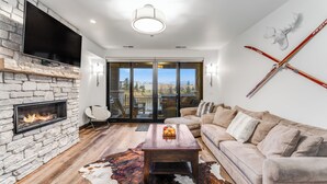 A modern living room with a stone fireplace, a flat-screen TV, a wooden coffee table with fruit, a beige sofa, and a large window with a view of the outdoors. - Hot Tub + Close to Downtown, Smith Fields and Whitefish Mountain Resort! (Whitefish)