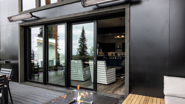 Un patio moderno al aire libre con una mesa de vidrio y una chimenea integrada, terraza de madera y vistas al interior a través de amplias puertas correderas de vidrio.