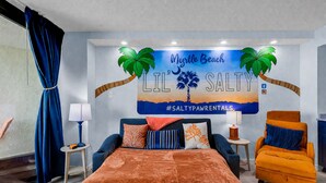 Una habitación de hotel con una cama, dos mesitas de noche, un sillón azul y otro naranja, y un mural con una escena de playa.