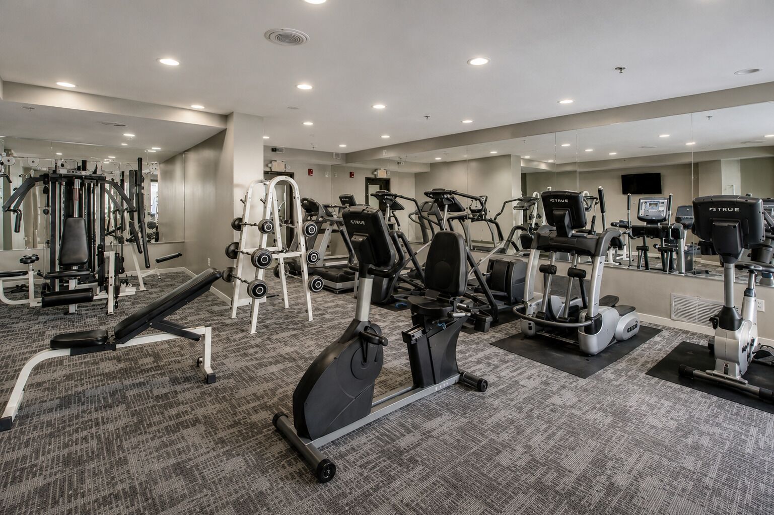Ein Fitnessraum mit verschiedenen Trainingsgeräten, Teppichböden und Spiegeln an den Wänden.