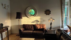 Un salon avec un canapé en cuir marron, une table basse en bois, un lampadaire, une fenêtre ronde donnant sur un paysage verdoyant et une petite table d’appoint avec un vase.