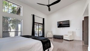 A bedroom with a bed, ceiling fan, TV, two chairs, and a balcony with a view of palm trees. - ScottsdaleStays 10BR-6BA Multimillion Acre Estate-Pool-SportCourts-SPECIAL DEALS (Phoenix)
