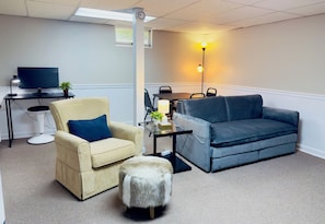 A living room with a beige sofa, a blue sofa, a small round ottoman, a TV, a lamp, and a plant.
