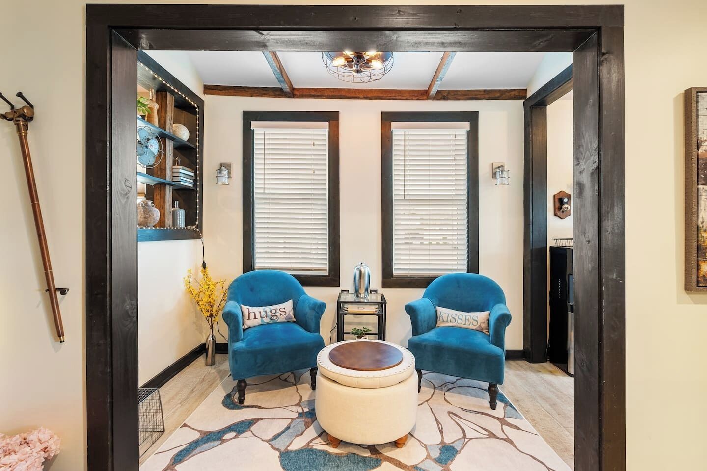 A room with two blue armchairs, a round ottoman, a bookshelf, and a chandelier.