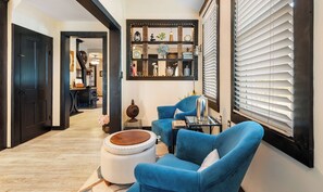 A hallway with a wooden door, a blue armchair, a round ottoman, and a shelf with various decorative items.