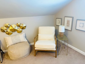 A room with a white armchair, a small ottoman with a patterned cushion, and a side table with a lamp.