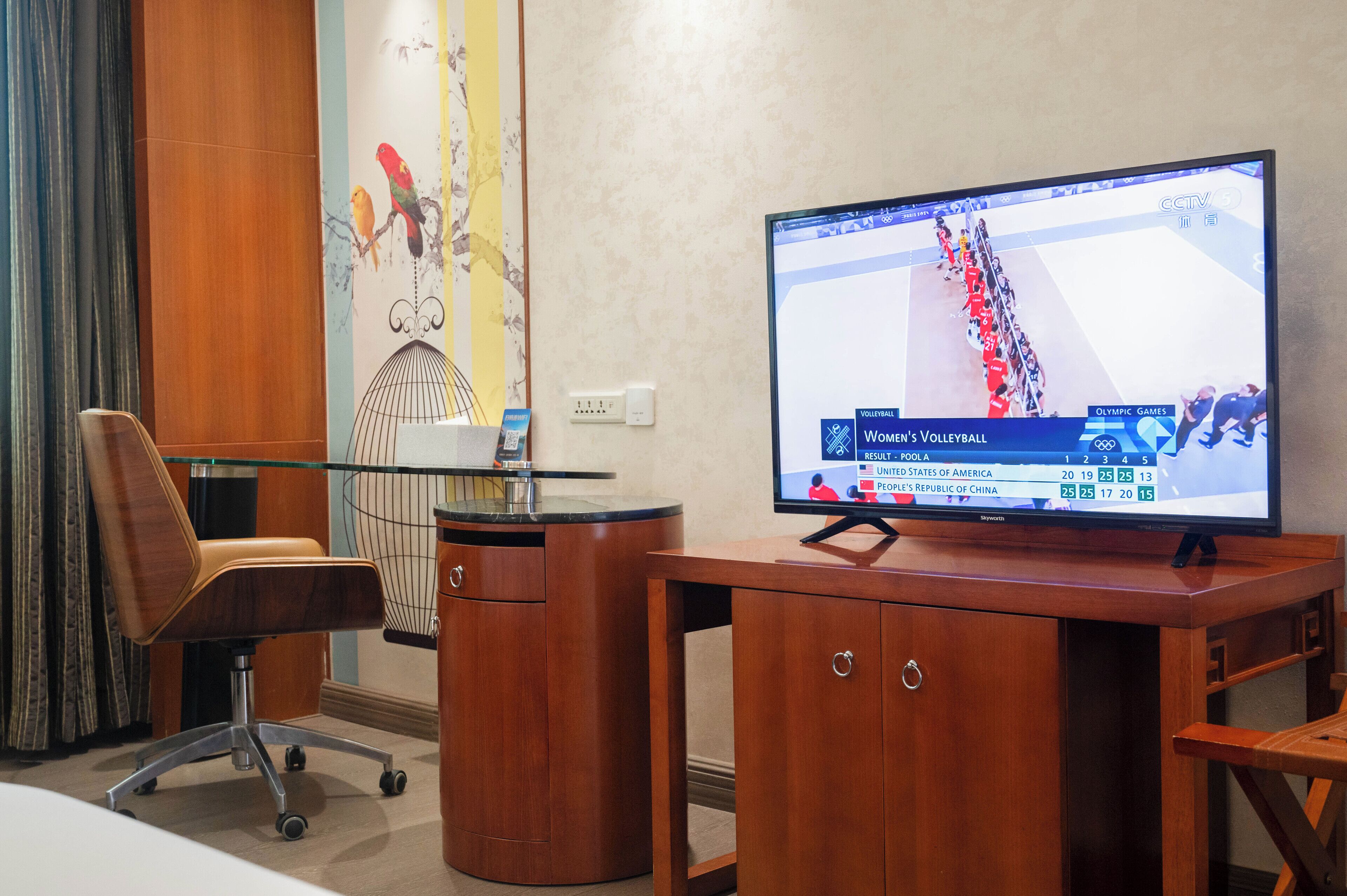 Une chambre d’hôtel avec un bureau en bois, un téléviseur diffusant les Jeux olympiques, une chaise et un mur décoratif.