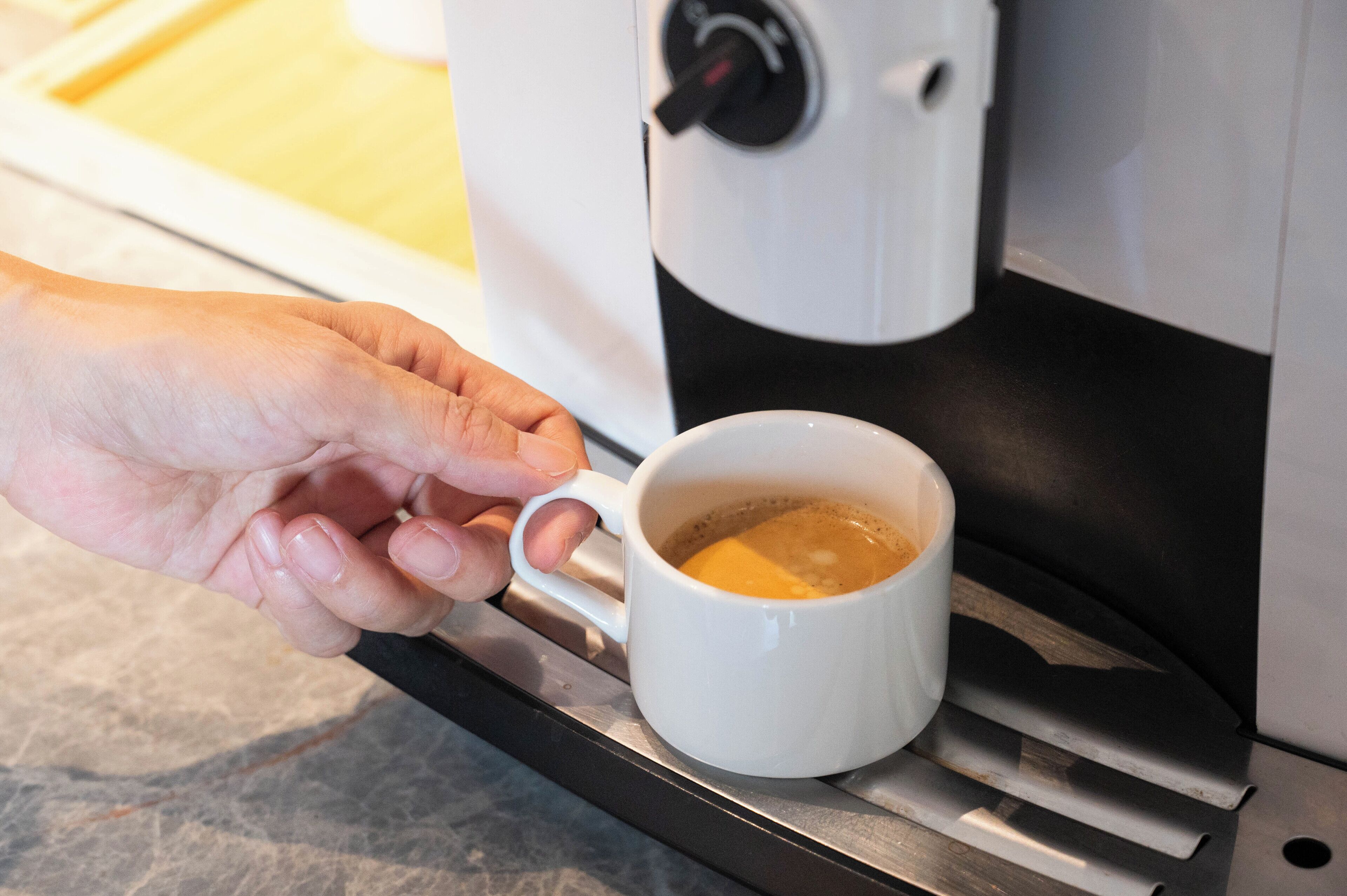 A hand holding a white mug filled with coffee, placed on a metallic tray next to a coffee machine.