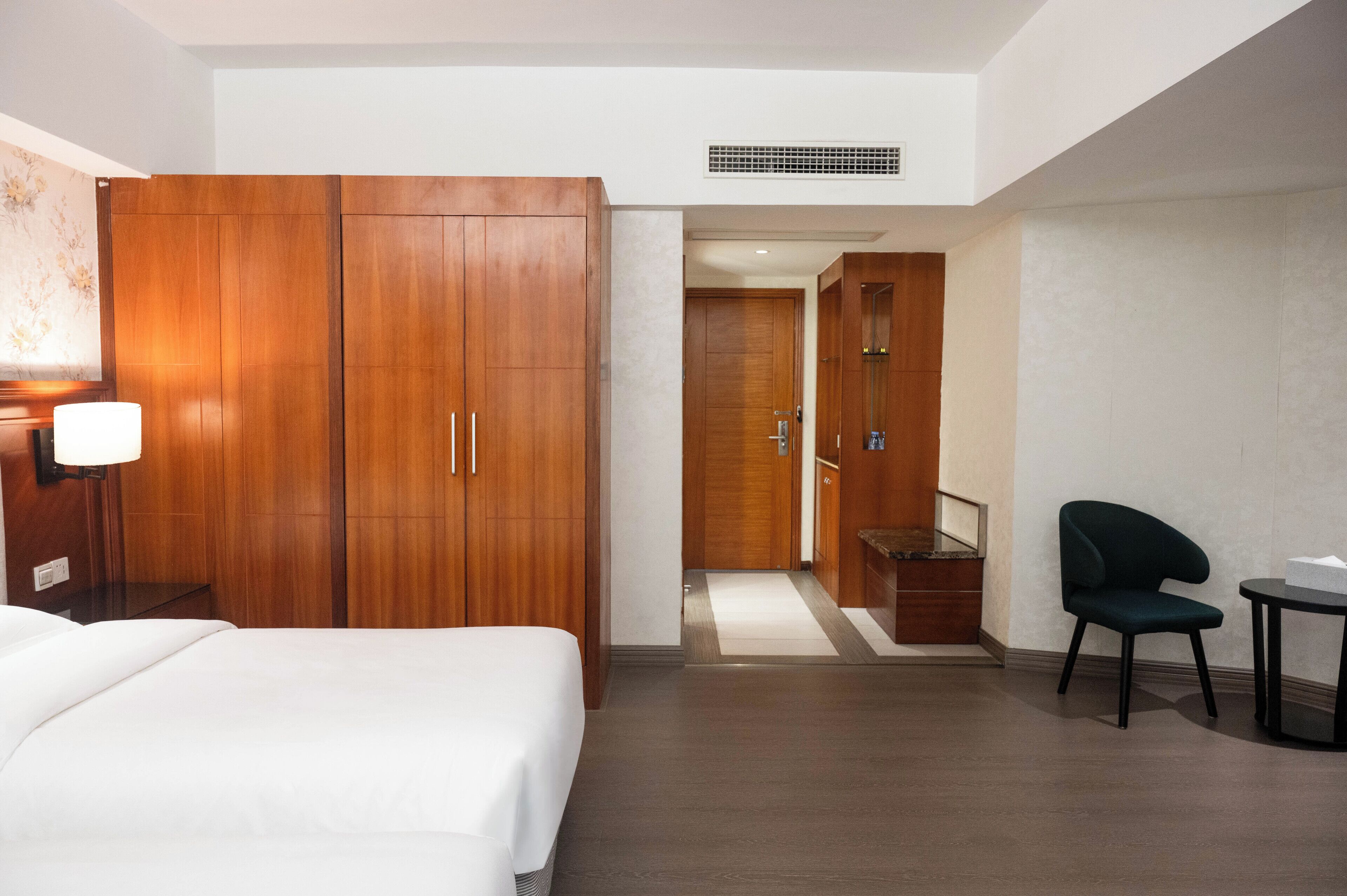 Una habitación de hotel con un armario de madera grande, una cama con ropa blanca, una mesita con lámpara y una silla.
