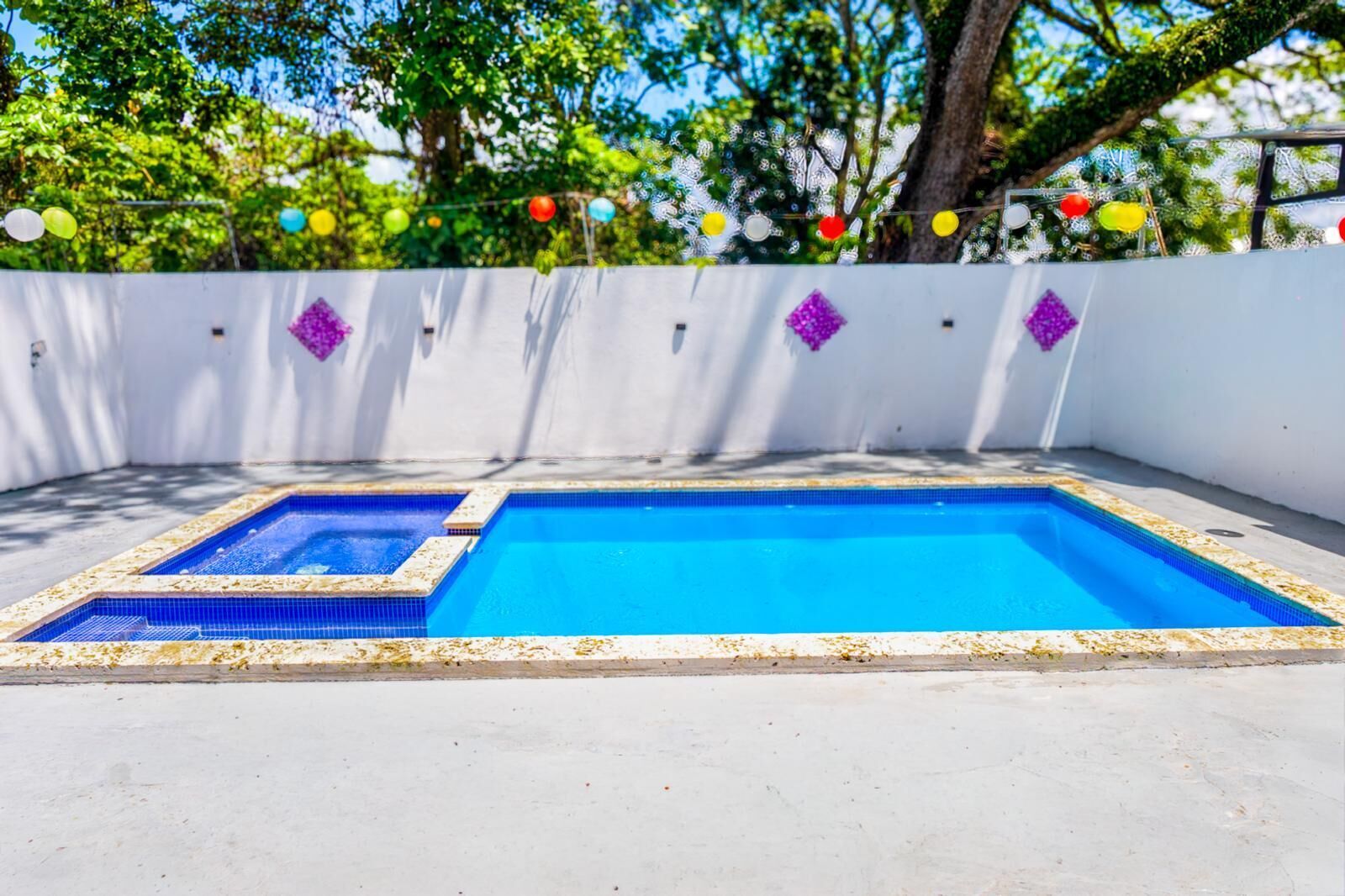 Piscina rectangular con un área pequeña adyacente, rodeada por un muro blanco con detalles decorativos morados.