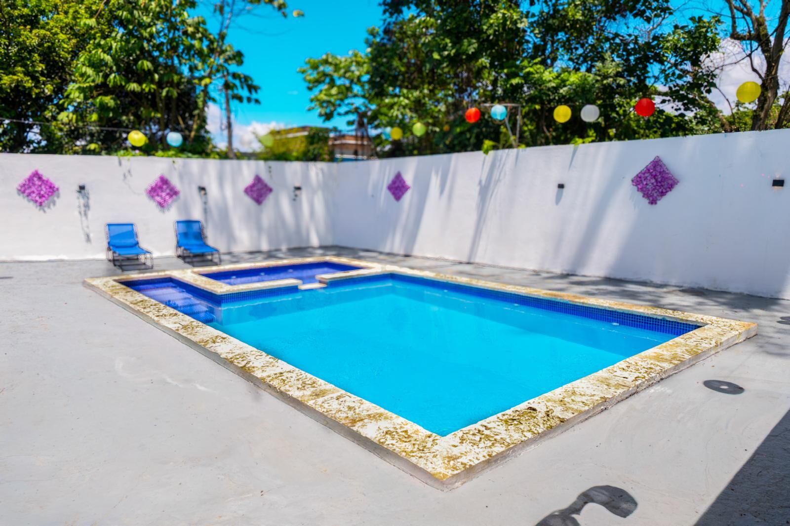 Una piscina rectangular con borde de azulejos, rodeada por una pared blanca con diseños de diamantes morados y sillas de descanso azules.