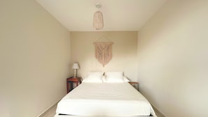 A bedroom with a bed, a hanging macrame wall decoration, a bedside table with a lamp, and a small wooden stool. - Fare Hinaiti - Premium Private Studio with sea vue and terrace (Faaa)